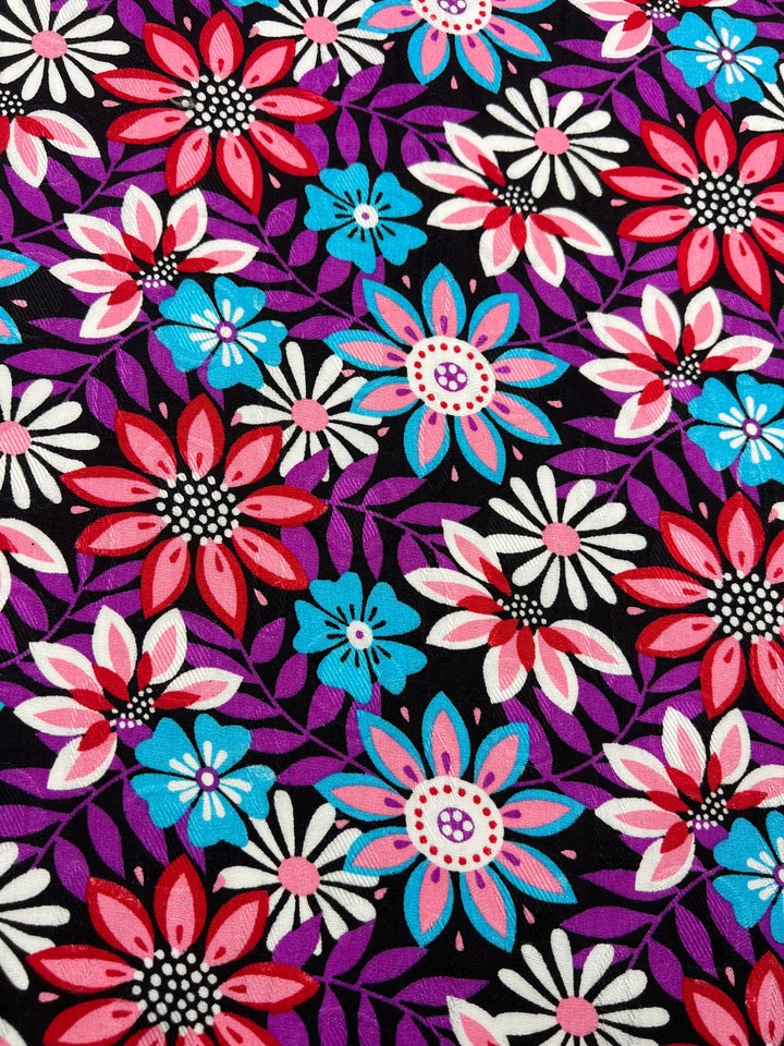 The Printed Rayon - Lolita Night from Super Cheap Fabrics features a vibrant floral pattern showcasing red, pink, blue, and white flowers against a black background. The flowers come in various sizes with detailed petals and centers, complemented by purple leaves for an intricate and colorful design. This 145cm wide fabric is ideal for creating versatile clothing with elegance.