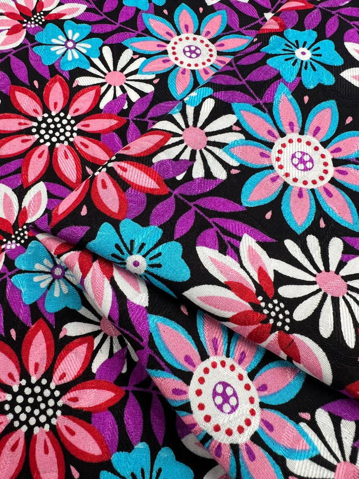 A close-up of the Printed Rayon - Lolita Night from Super Cheap Fabrics, showcasing a vibrant floral pattern. The design features large, bright flowers in shades of red, pink, blue, white, and purple with intricate details set against a black background. This versatile clothing material is slightly folded at the bottom and measures 145cm in width.