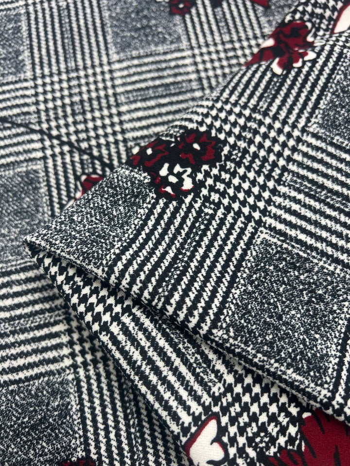 A close-up of the Scuba Crepe - Hounds Dream - 160cm from Super Cheap Fabrics, showcasing a classic black-and-white houndstooth and checkered pattern adorned with scattered red and white floral accents. The edges of the textured, lightweight fabric are folded to highlight its intricate design details.