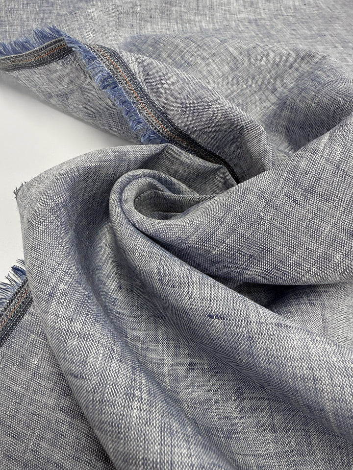 A close-up of Pure Linen - Indigo Husk - 135cm by Super Cheap Fabrics, featuring textured indigo linen with a visible weave and frayed edges, loosely gathered and slightly twisted on a white surface.
