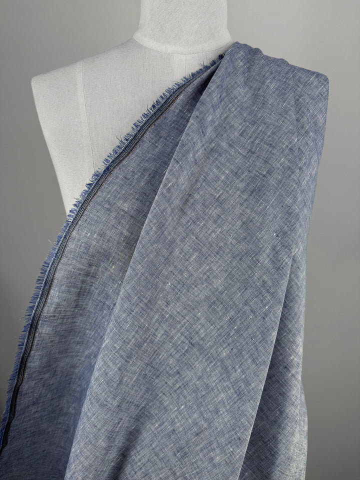 A mannequin displays Super Cheap Fabrics' Pure Linen - Indigo Husk (135cm), a soft, lightweight blue linen fabric with a raw fringed edge, set against a plain gray background.