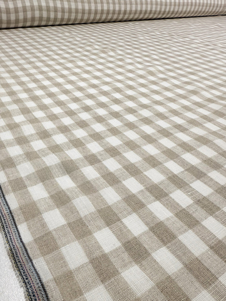 A bolt of Super Cheap Fabrics' Pure Linen - Oatfield Check (140cm), featuring a beige and white gingham checkered pattern, is rolled out to showcase its 100% linen texture and repeating squares.