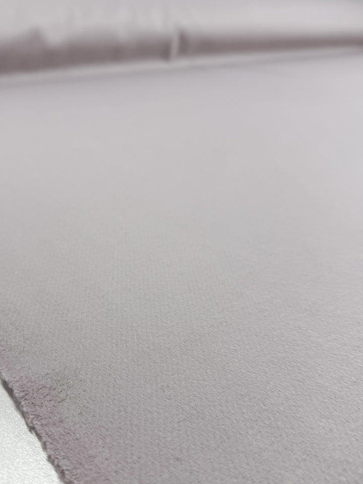 A close-up of Super Cheap Fabrics' Wool Cashmere - Windchime (150cm) shows a smooth, light gray surface with subtle texture, a visible edge in the foreground, and a softly blurred background.