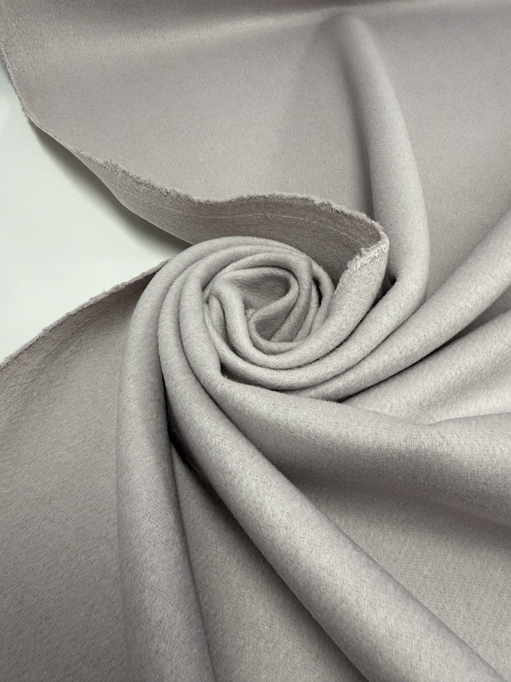 A close-up of Super Cheap Fabrics’ Wool Cashmere - Windchime - 150cm shows its light gray, soft, and smooth premium fabric arranged in a spiral with visible raw edges.
