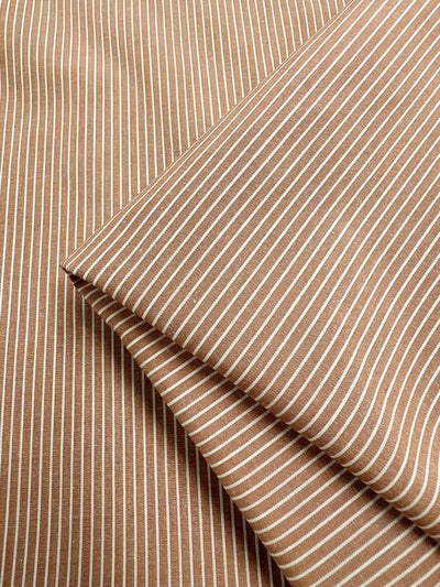 Close-up of the Super Cheap Fabrics' Shirting - Marmalade, a brown polyester cotton blend with thin white vertical pinstripes, neatly folded. This fabric offers a smooth and lightweight texture with evenly spaced stripes that create a classic and elegant pattern. The width of the fabric is 147cm.