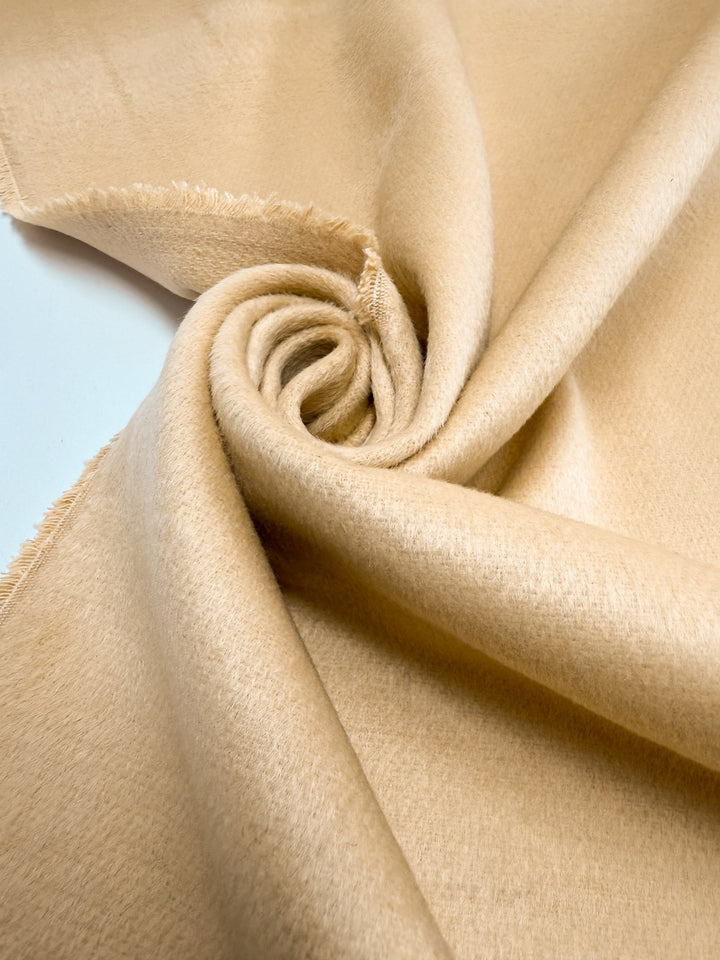 A close-up of Super Cheap Fabrics' Wool Cashmere - Banana Crepe (150cm) fabric in light beige, showing its soft texture and frayed edges spiraled into a swirl.