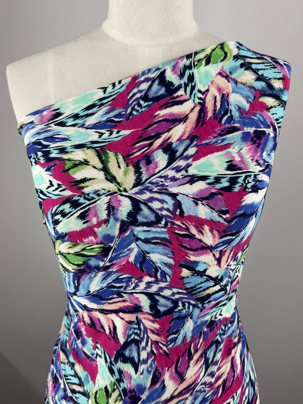 A mannequin sports a vibrant dress in medium-weight Printed Lycra by Super Cheap Fabrics, adorned with bold, feather-like abstract patterns in pink, blue, green, and black. The Polyester/Spandex blend offers stretch to the one-shoulder design with an asymmetrical neckline.