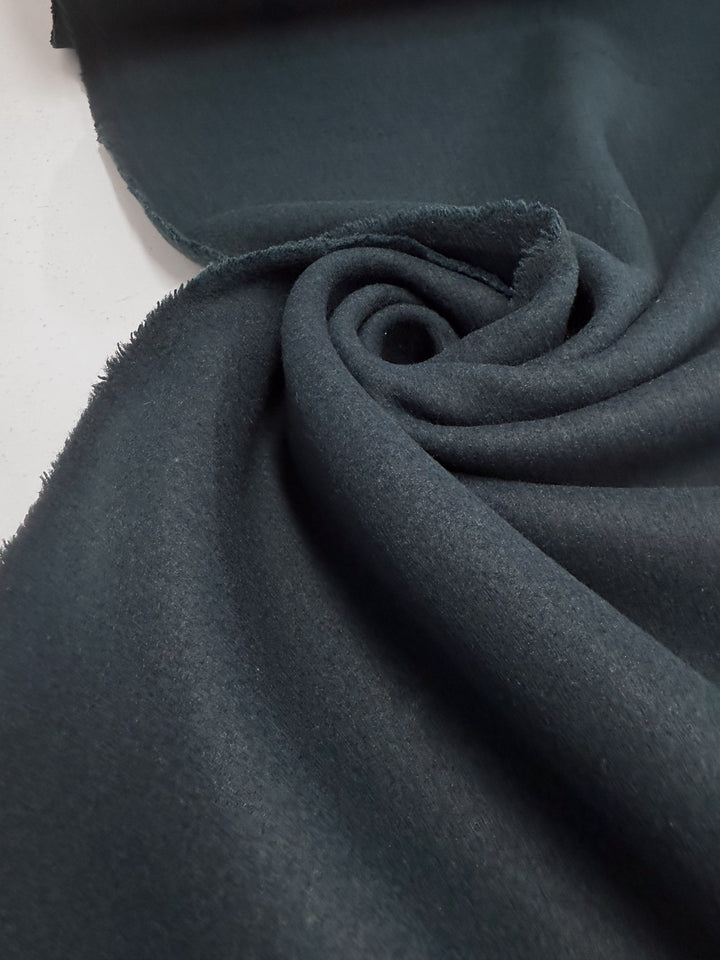 A close-up of Super Cheap Fabrics’ Wool Cashmere - Gulf Coast (154cm): dark gray, softly textured fabric with one edge rolled in a spiral and the other frayed, shown on a light surface.