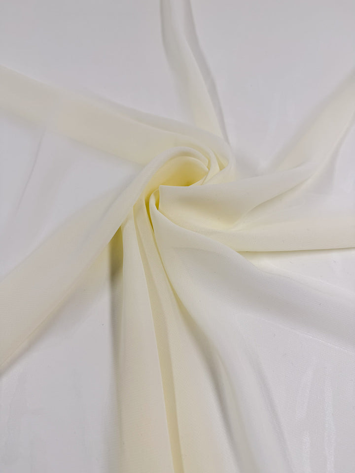 The Hi-Multi Chiffon - Cream - 150cm from Super Cheap Fabrics is displayed on a white surface, gathered and swirled at the center with soft folds of sheer polyester radiating outward.
