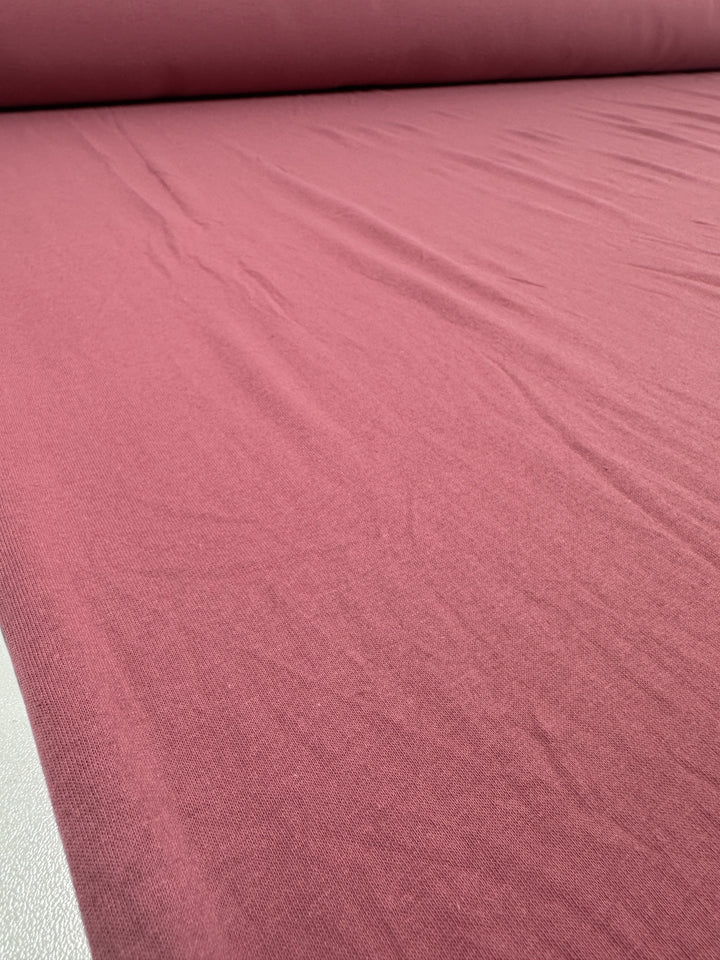 A close-up of a large roll of Super Cheap Fabrics’ Cotton Jersey in Slate Rose, 175cm wide, showcases its smooth texture with subtle creases. Gentle light reflection suggests the soft and possibly stretchy nature of this pink-hued fabric.