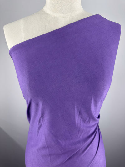 A mannequin showcases Super Cheap Fabrics' Cotton Jersey in Patrician Purple, an off-the-shoulder fabric draped diagonally with soft texture and elegant folds against a simple gray background.