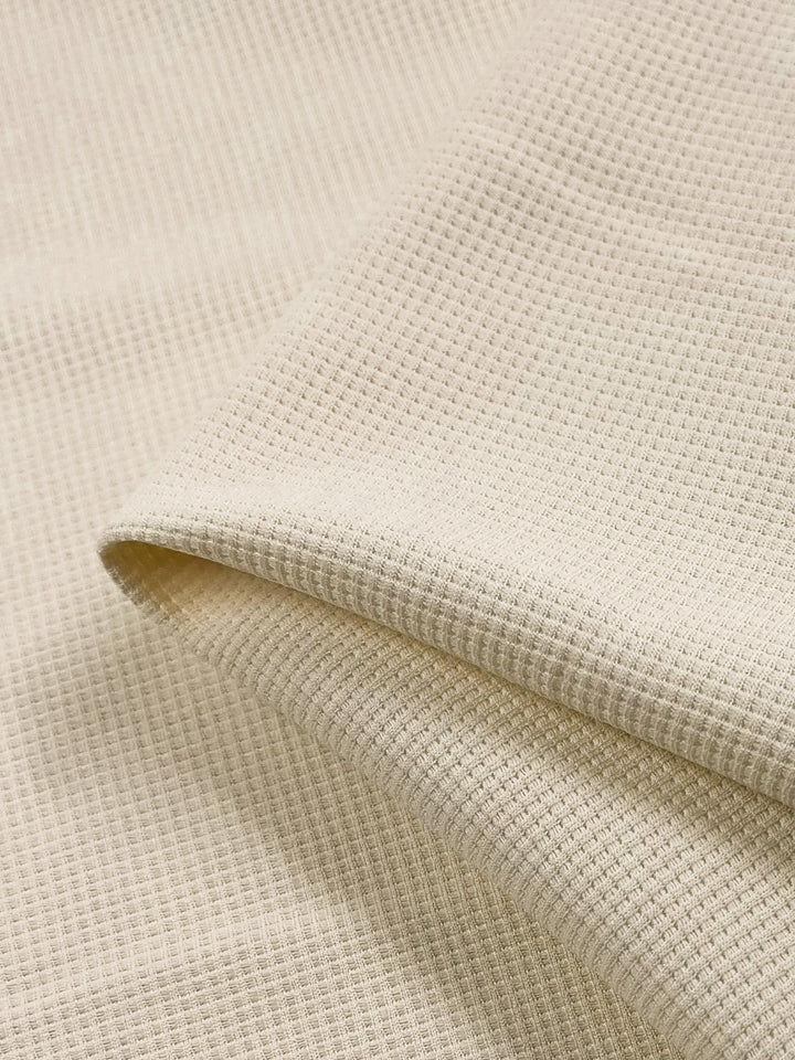 A close-up image showcases the folded texture of Super Cheap Fabrics' "Waffle Knit - Cream - 170cm," characterized by a three-dimensional appearance. The soft and inviting look is created by small, evenly spaced square patterns in the weave, resulting in a smoothly draped fabric with visible details.