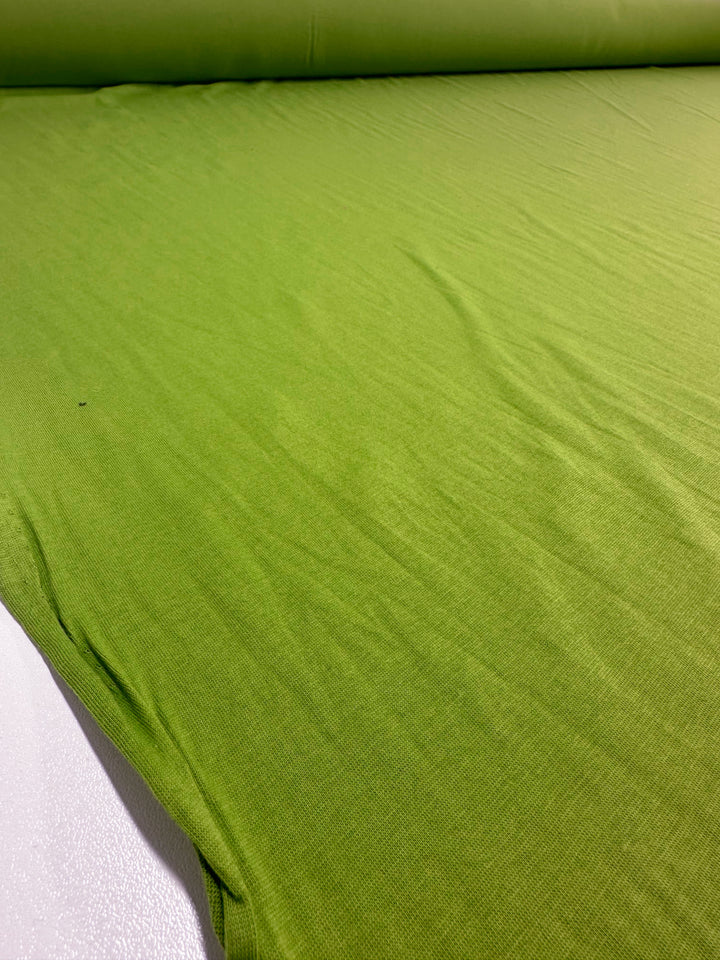 A large roll of Super Cheap Fabrics' Cotton Jersey - Titanite - 160cm in vivid green is unrolled on a flat surface, featuring a subtle texture and slight ripples that highlight its soft and flexible cotton blend.