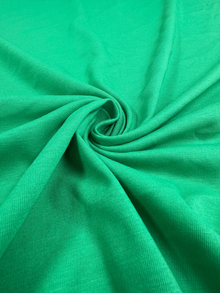 Close-up of Super Cheap Fabrics' Cotton Jersey in Vivid Green, 165cm, with a smooth texture displayed in a swirl pattern, showcasing its soft folds and rich color.