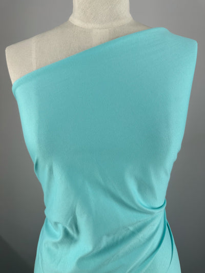 A mannequin displays Super Cheap Fabrics' Cotton Jersey in Aqua Splash - 160cm, elegantly draped to create gentle folds. The fabric cascades from one shoulder against a plain grey background, highlighting its soft allure.