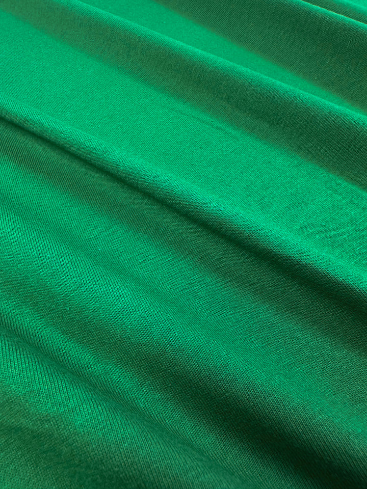 Close-up of Super Cheap Fabrics' Cotton Jersey in Abundant Green, 165cm. This 100% cotton material showcases visible texture and folds with subtle diagonal lines, adding depth and movement.