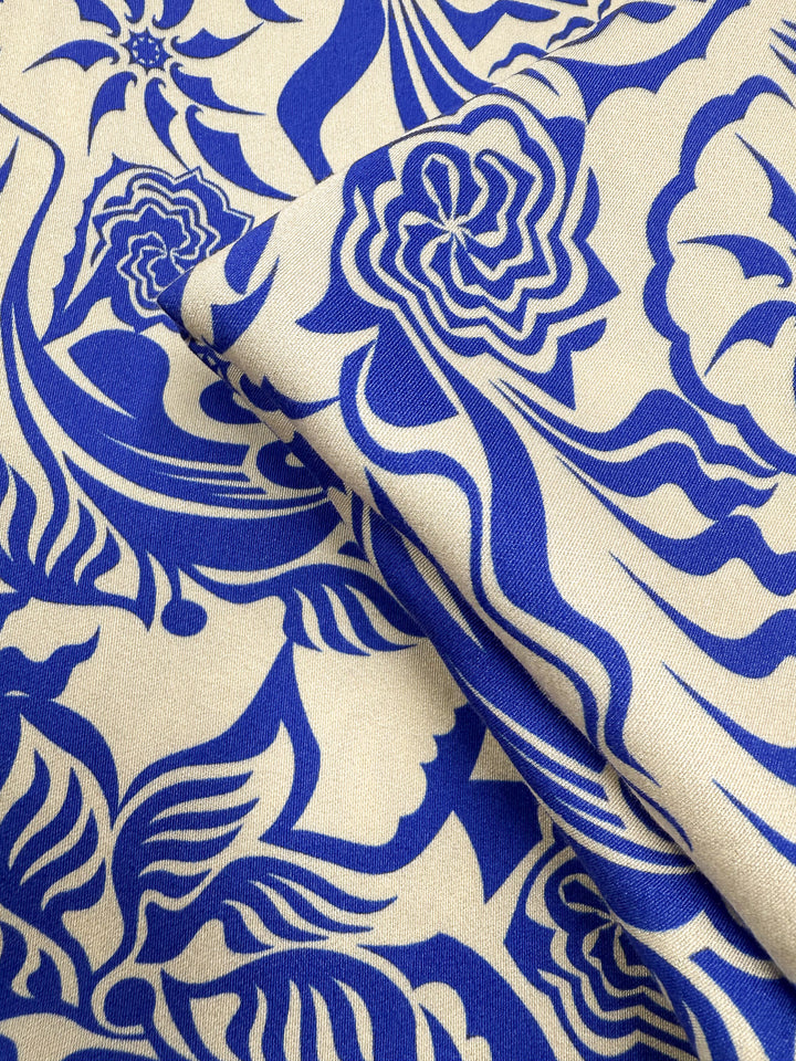 A close-up of Mercerised Rayon - Cobalt Whirl (150cm) from Super Cheap Fabrics shows breathable fabric with a detailed blue floral and abstract pattern on cream, featuring stylized flowers, leaves, and flowing lines.