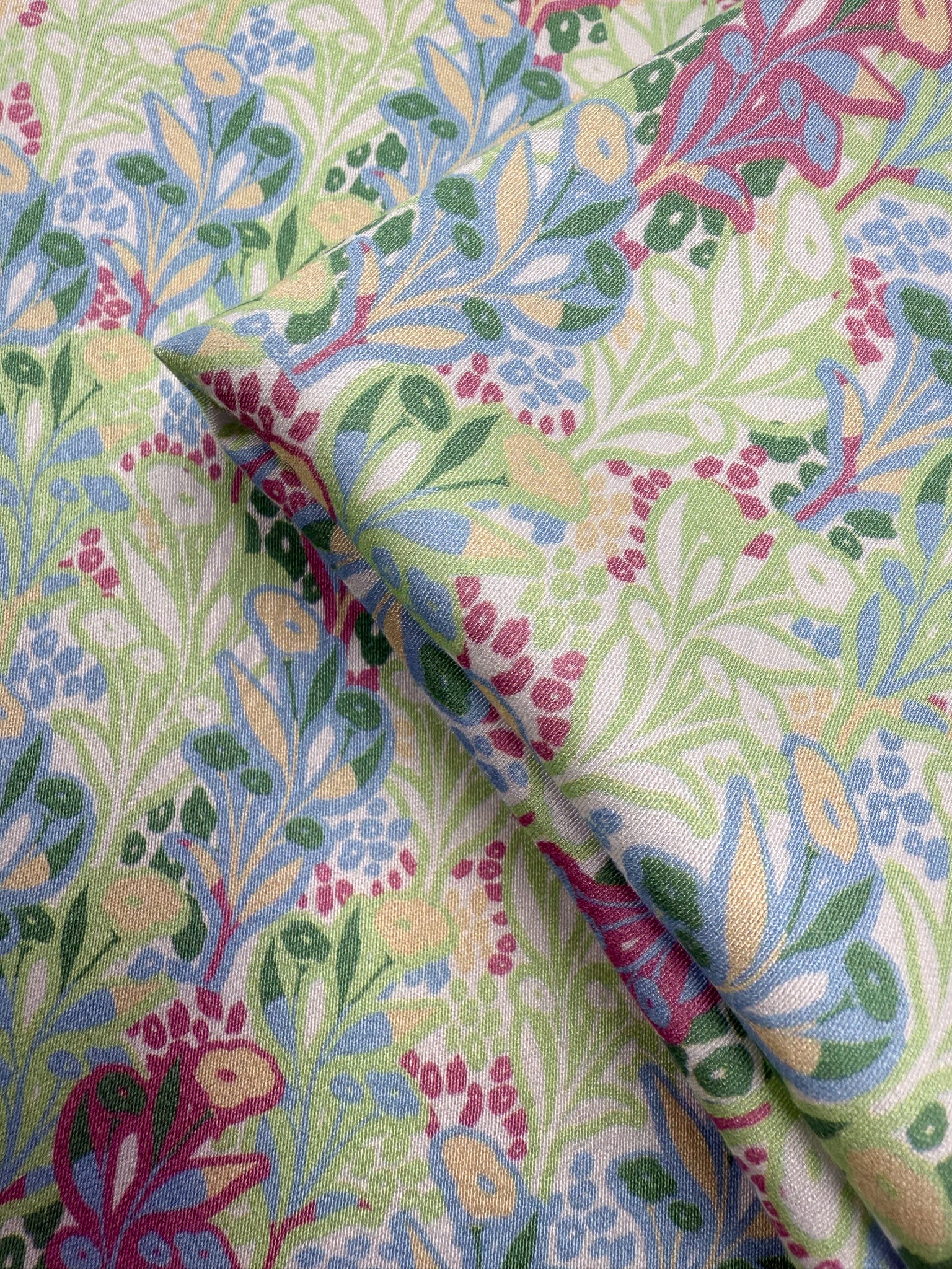Close-up of Mercerised Rayon - Garden Vine (150cm) by Super Cheap Fabrics, featuring breathable rayon with a colorful floral pattern of green leaves, blue and yellow flowers, and red berries on a light background.