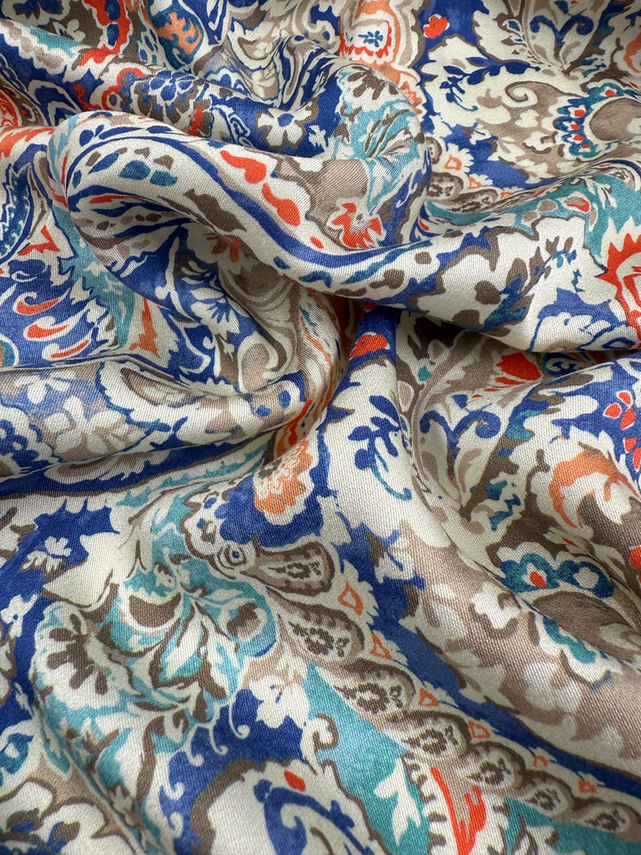 A close-up of Super Cheap Fabrics' Mercerised Rayon - Paisley Fusion (150cm), featuring lightweight rayon with a smooth, slightly glossy finish and a detailed blue, teal, red, and tan paisley on beige, softly draped and loosely gathered.