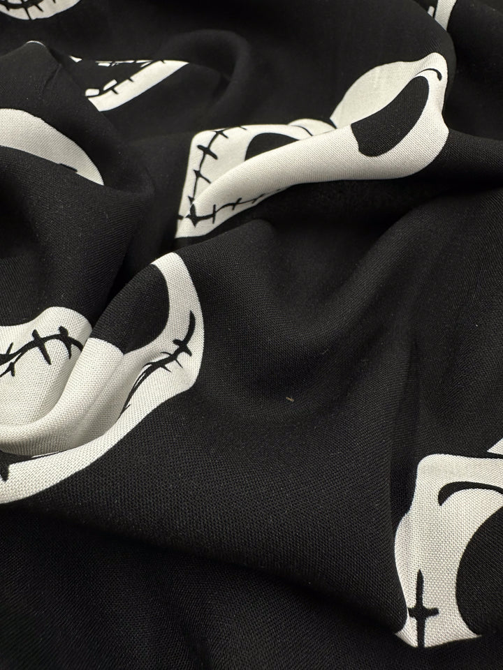 The Super Cheap Fabrics "Printed Rayon - Skellington - 140cm" showcases a close-up of black fabric with white skulls and stitched outlines. Its soft, slightly crumpled look adds shadow and texture for versatility.