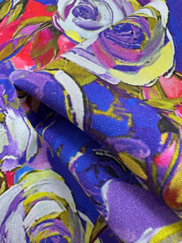 A close-up of the Designer Rayon - Zoe - 145cm by Super Cheap Fabrics showcases vibrant purple, pink, yellow, and blue roses. The fabric's folds highlight its vivid colors and intricate design—a versatile choice for adding vibrant prints to any project.