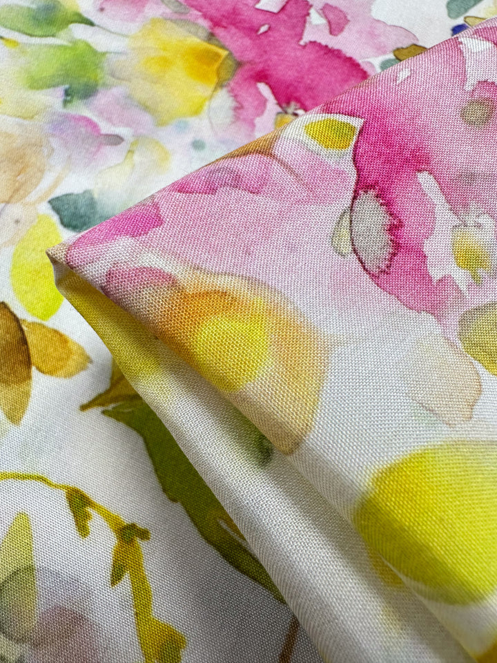 A close-up of Designer Rayon - Jesse by Super Cheap Fabrics displays a watercolor floral pattern with pink, yellow, and green flowers on white. The soft fabric is gently folded, showcasing vibrant prints in an artistic style.