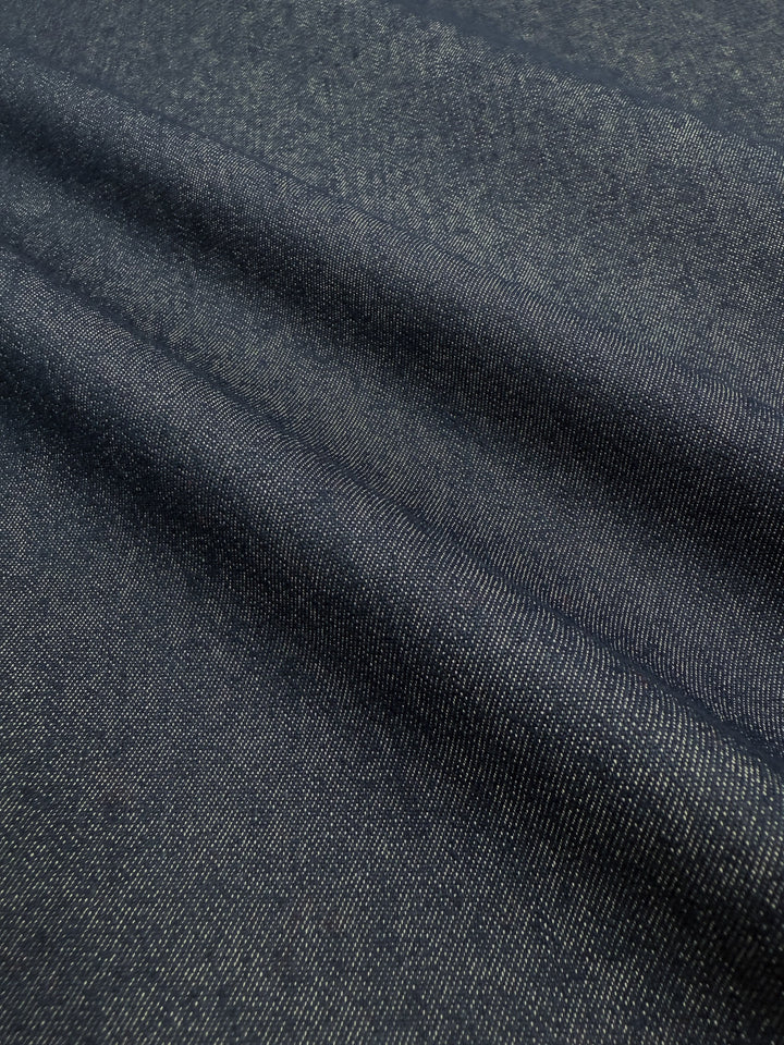 Super Cheap Fabrics’ Denim - Classic - 150cm is a dark blue 100% cotton fabric featuring diagonal ridges and a textured weave, shown under soft lighting to highlight its texture and subtle sheen.