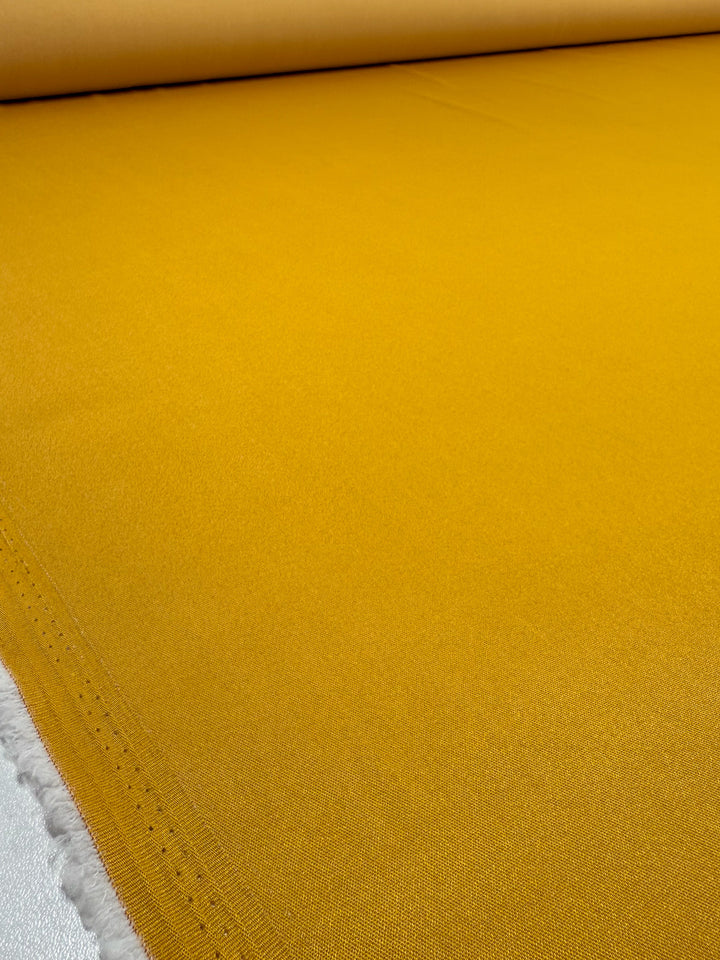 A close-up of Super Cheap Fabrics’ Stretch Denim - Mustard - 150cm shows the vibrant mustard fabric roll neatly spread out with its selvage edge visible on the left.