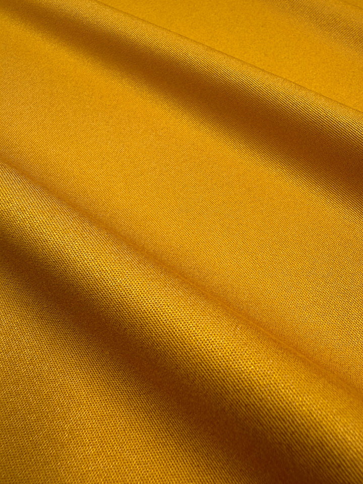 Close-up of Super Cheap Fabrics' Stretch Denim - Mustard - 150cm: smooth, slightly shiny surface with gentle diagonal folds. The medium weight fabric appears soft, vibrant, and catches the light, highlighting its rich mustard color.