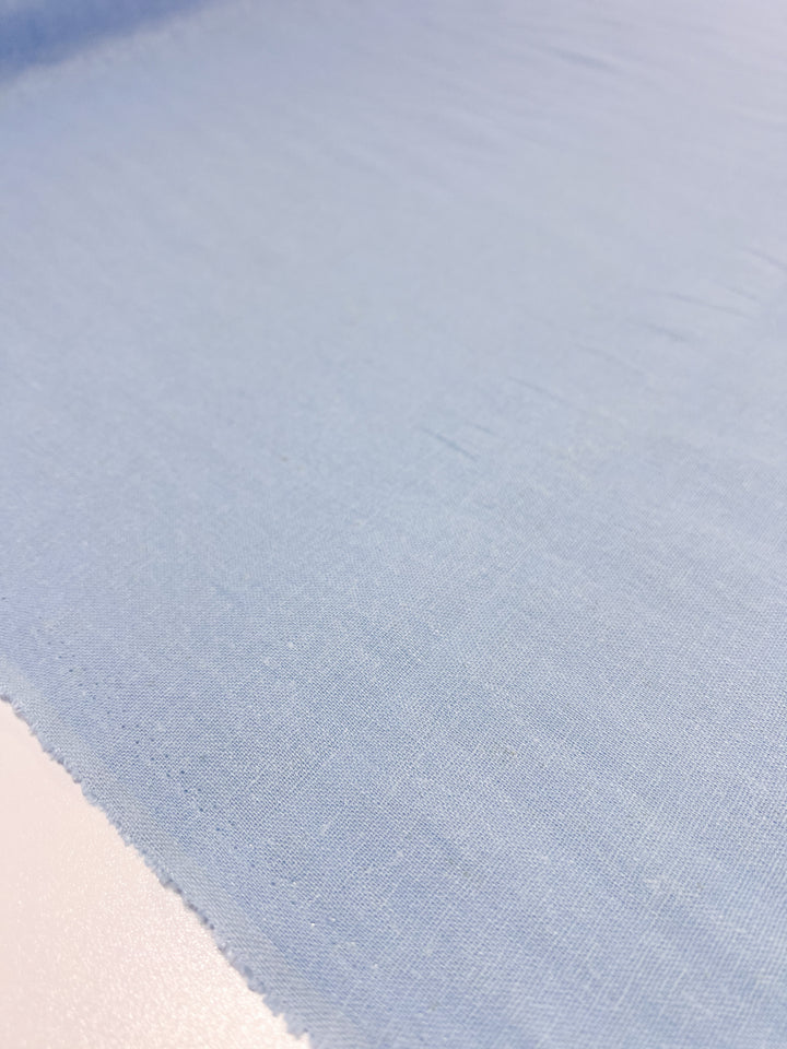 A close-up of the Premium Linen Cotton - Clear Sky fabric from Super Cheap Fabrics showcases its light blue, slightly textured surface, reminiscent of a clear sky, as it is spread out on a flat, white table. The evenly colored, lightweight material appears smooth and extends off into the distance.