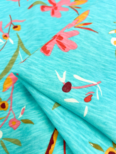 Close-up of the Printed Jersey - Evelyn by Super Cheap Fabrics, showcasing a teal poly/cotton blend adorned with a vibrant floral pattern. The design features pink and red flowers paired with leaves in shades of green and orange, offering a lively, colorful look ideal for printed jersey fabric enthusiasts.