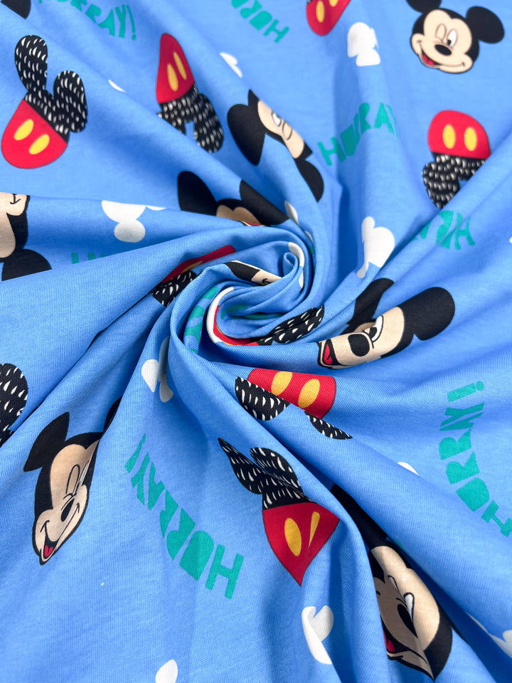 A playful two-way stretch Printed Jersey from Super Cheap Fabrics, featuring cartoon mouse faces with red pants and the word "Hooray!" in green and white. This medium-weight blue fabric is swirled to beautifully display its whimsical pattern, with a width of 170cm.