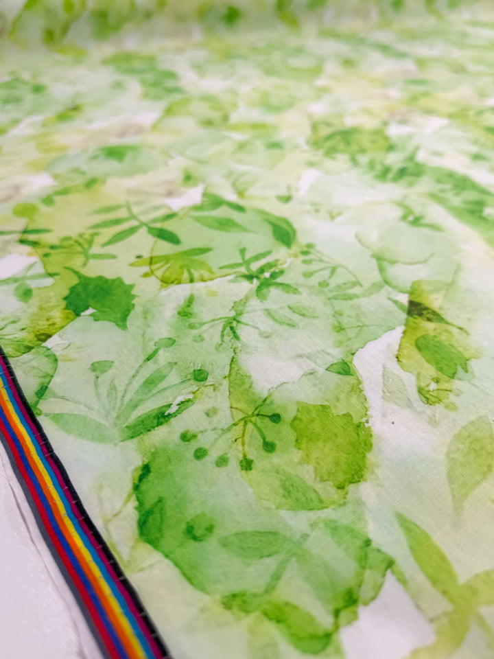 A close-up of Super Cheap Fabrics' Designer Cotton - Garden Fade (145cm), showcasing lightweight cotton with a green leaf and foliage print on white, plus a rainbow-striped edge on the left.