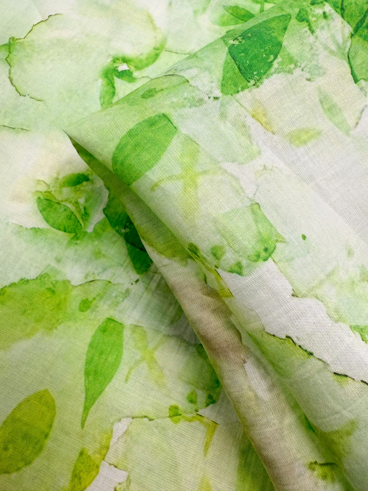 A close-up of Designer Cotton - Garden Fade by Super Cheap Fabrics shows a soft, lightweight cotton with a watercolor green leaf pattern on white, gently folded to highlight its texture—ideal for luxury sewing projects. Width: 145cm.