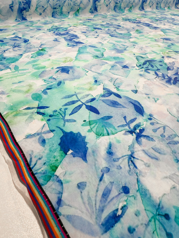 A close-up of Super Cheap Fabrics' Designer Cotton - Oceans (145cm), showcasing lightweight cotton with a blue and green watercolor leaf pattern and a colorful striped border—ideal for sewing projects, displayed flat.