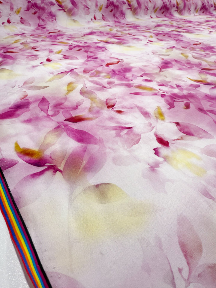 A close-up of Super Cheap Fabrics' Designer Cotton - Bloom Drift (145cm), a lightweight cotton featuring a pink, purple, and yellow floral print with soft watercolor-like petals and a thin multi-colored rainbow stripe along the selvage edge.