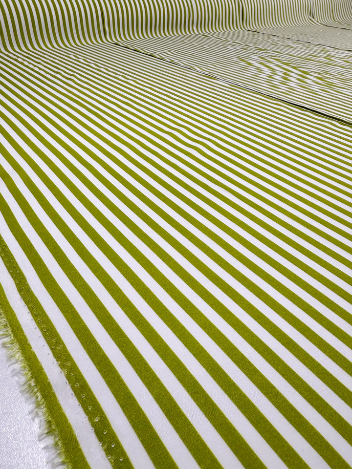 Super Cheap Fabrics’ Printed Rayon - Lime Pop Stripe - 140cm features yellow-green and white stripes that flow smoothly across the fabric, curving upward with a frayed edge visible on the left side for added texture.
