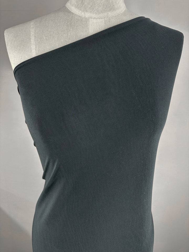 A mannequin is adorned with ITY Knit fabric in a charcoal shade from Super Cheap Fabrics, draped elegantly over one shoulder to achieve an asymmetrical style. The plain gray background accentuates the sophisticated simplicity of this medium weight fabric and its draping technique.