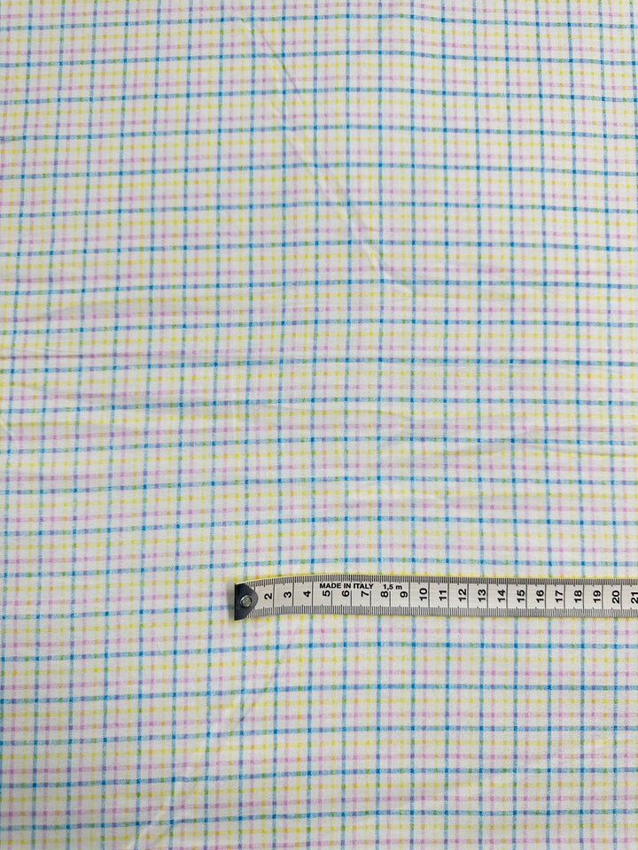 A lightweight, multi-colour white fabric with a small checkered pattern in blue, yellow, and green lines is displayed. A measuring tape laid across the bottom of the Super Cheap Fabrics Flannelette - Tattersall - 110cm shows a length in centimeters, ranging from 10 to 27, indicating the scale of the pattern.