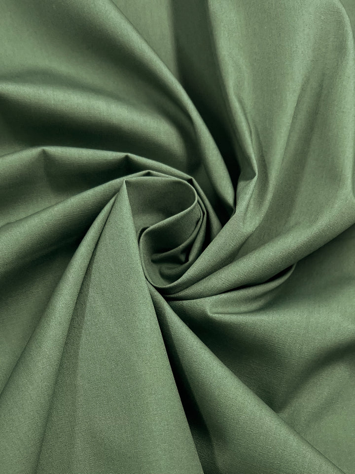 The Poplin - Silver Pine fabric by Super Cheap Fabrics features a lightweight polyester cotton blend with a gently textured surface, artfully folded into swirling pleats to form a central spiral pattern. Its smooth, silky finish reflects light softly. Width: 148cm.