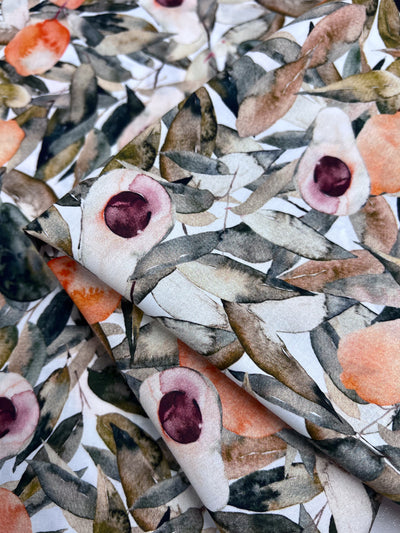 A close-up of Super Cheap Fabrics' Printed Cotton - Avo Tree - 150cm shows its lightweight material with watercolor florals, leafy green branches, and abstract flowers in peach, pink, and purple on white. The fabric is folded to reveal its texture and natural cotton fibers.