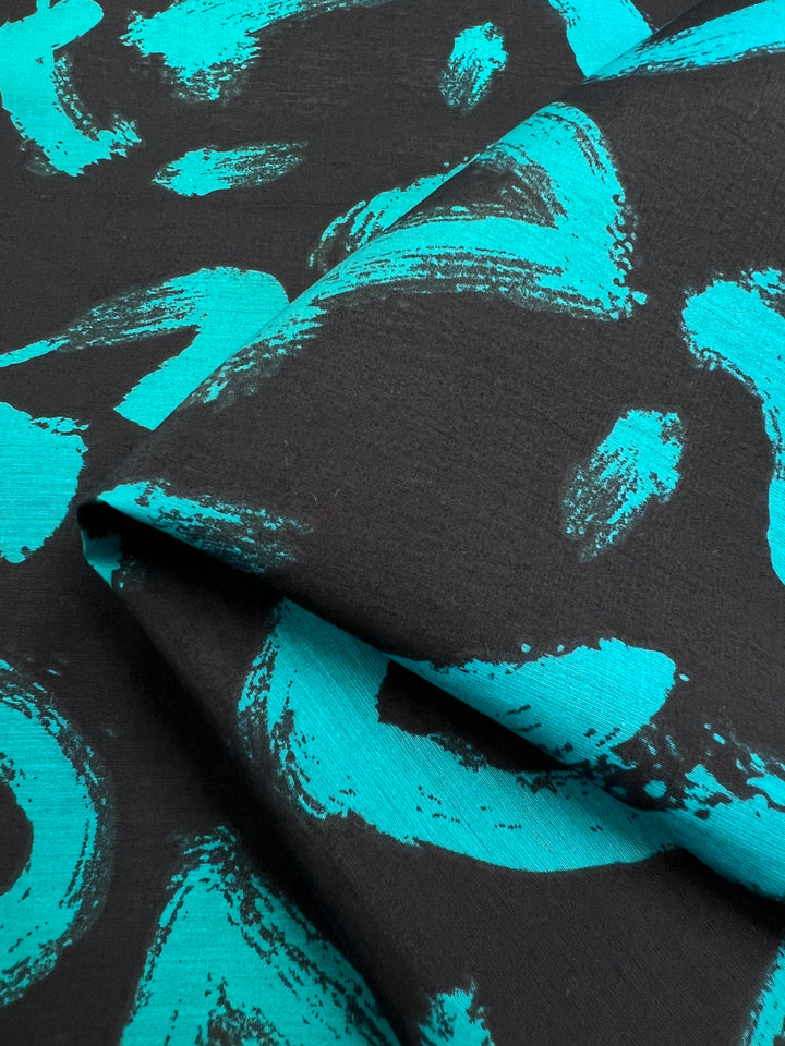 The Printed Cotton - Texter from Super Cheap Fabrics features a dynamic turquoise abstract pattern on lightweight fabric, with brushstroke-like designs on a black background. This 150cm versatile fabric is perfect for stylish and creative projects.