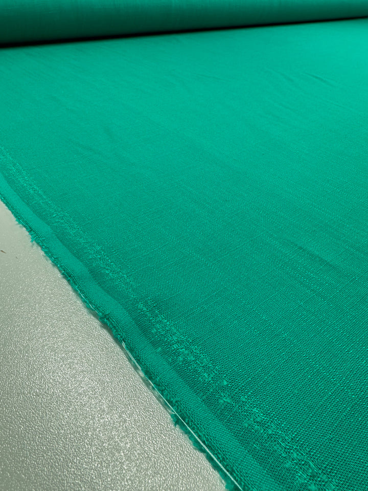 A close-up of a roll of Linen Blend - Sea Green - 135cm by Super Cheap Fabrics, unfurled on a flat surface. The fabric shows a smooth, even texture with a slightly frayed edge on one side. The surface underneath the Linen Blend has a slight shimmer, reflecting light softly.