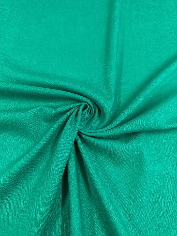 A close-up of a piece of sea green fabric gathered into soft folds and wrinkles, creating a swirling pattern at the center. The texture appears smooth and slightly shiny, indicating that the material might be Super Cheap Fabrics' Linen Blend - Sea Green - 135cm, perfect for table linens.