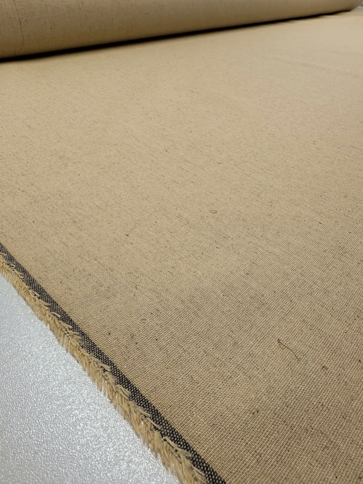 A close-up view of a beige Linen Blend - Short Bread - 135cm roll from Super Cheap Fabrics with visible texture and a black-bordered edge, placed on a smooth, light-colored surface. The fabric appears to have a nubby texture and is tightly rolled at one end, perfect for creating elegant Table Linens.