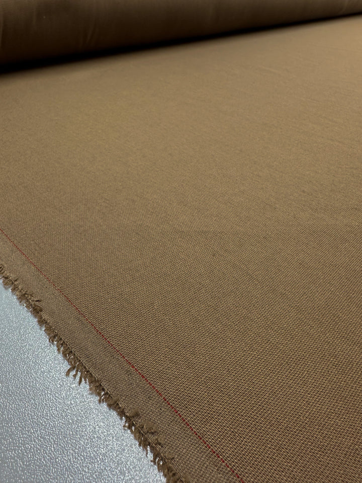 Close-up of a large roll of Linen Blend - Cocha Mocha - 137cm by Super Cheap Fabrics with a visible selvage edge. The fabric is unrolled over a flat surface, displaying its texture and weave. One edge shows a frayed finish with a faint red line running parallel to it, perfect for crafting table linens.