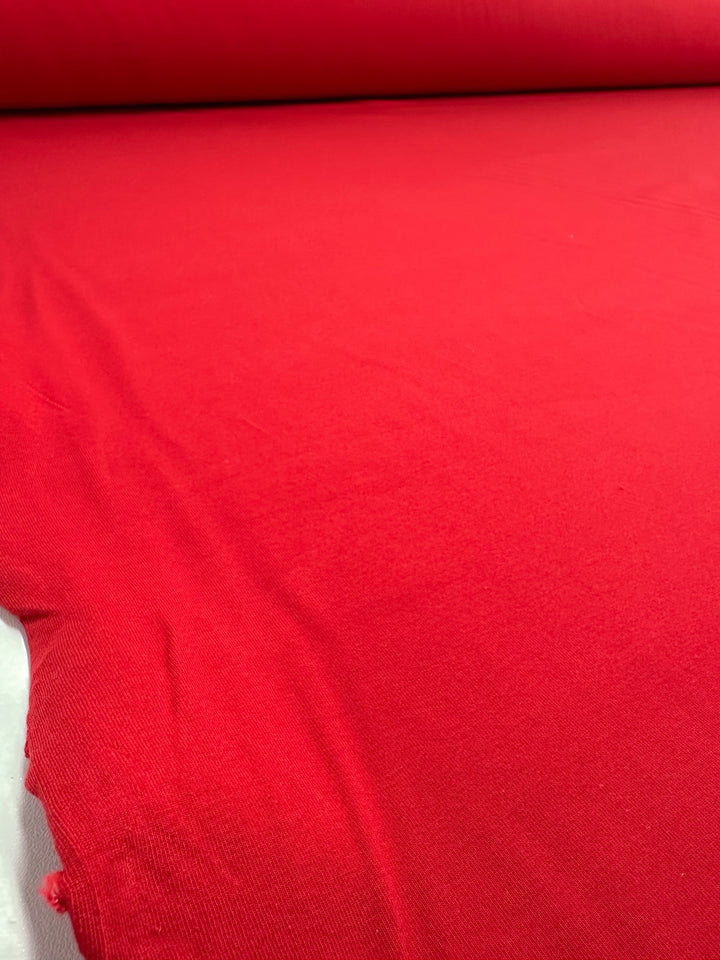 A vivid close-up showcases a large, smooth red Cotton Lycra fabric from Super Cheap Fabrics. This Goji Berry-hued medium-weight material is slightly textured with minor wrinkles and features a rolled edge, ideal for Bodycon dresses. Fabric width measures 170cm.