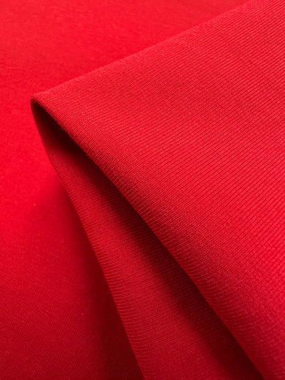 Close-up of Super Cheap Fabrics' Cotton Lycra in Goji Berry (170cm), showcasing its bright red hue and smooth texture. The fabric is folded with a sharp triangular crease, highlighting the subtle weave pattern—ideal for crafting stylish bodycon dresses.