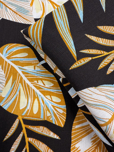Close-up of Super Cheap Fabrics' Bamboo Rayon - Tropic Gold (150cm), featuring a tropical leaf pattern in mustard yellow, white, and light blue on dark fabric; two angled layers—ideal for beginner sewing projects.