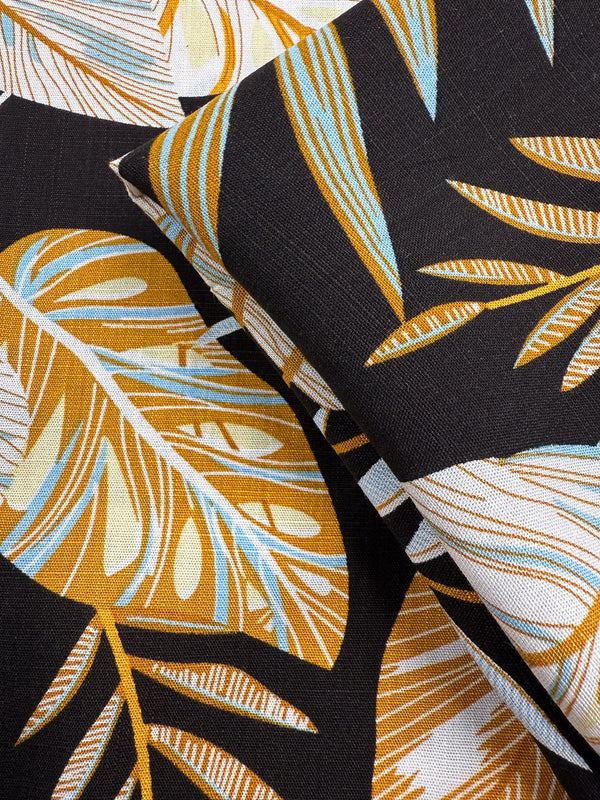 Close-up of Super Cheap Fabrics' Bamboo Rayon - Tropic Gold (150cm), featuring a tropical leaf pattern in mustard yellow, white, and light blue on dark fabric; two angled layers—ideal for beginner sewing projects.